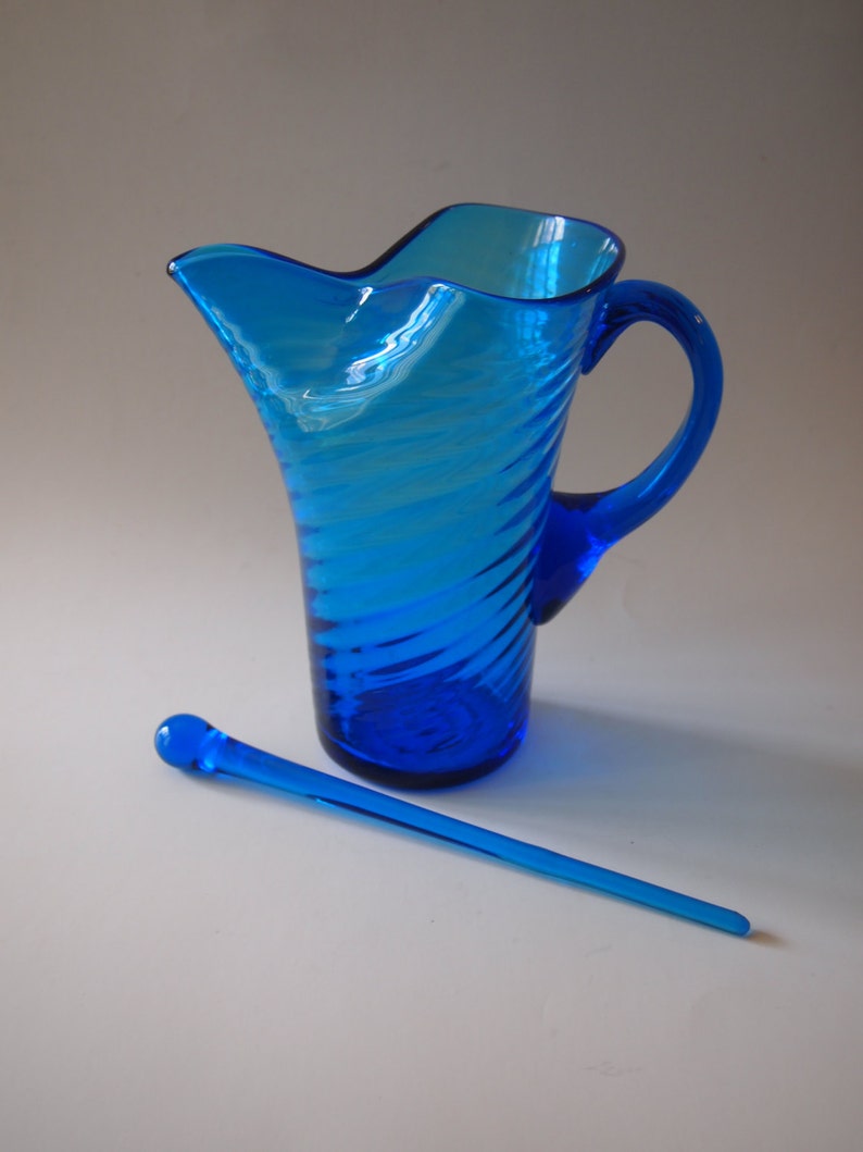 May include: A vibrant blue glass pitcher with a spiral design and a matching stirrer. The pitcher has a curved handle and a unique, wavy spout. The stirrer is long and slender, with a rounded end. The items are displayed on a white surface.