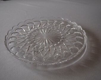 May include: A clear, decorative glass plate with a scalloped edge. The plate features a detailed, repeating pattern of diamond and teardrop shapes radiating from the center. The plate is designed to be used for serving food.