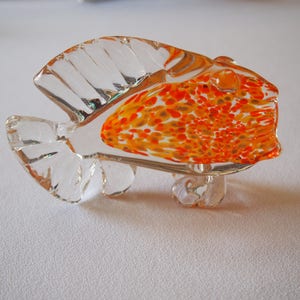 May include: A clear glass fish figurine with orange and yellow speckled detailing. The fish has a detailed fin design and is resting on a clear glass base. The figurine is a decorative item.