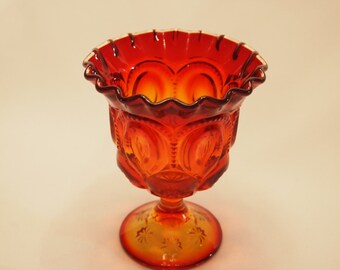May include: A vintage, orange-red glass pedestal bowl. The bowl has a ruffled rim and a detailed pattern of crescent moons and stars. The base is round with a starburst design. The glass has a glossy finish.