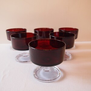 May include: Six ruby red glass dessert bowls with clear stems and bases are arranged on a white surface. The bowls have a rounded shape and a deep red color, perfect for serving desserts or small portions.