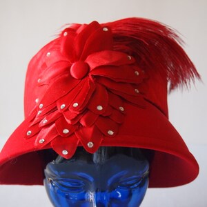 May include: A vibrant red hat with a decorative flower and feather accents. The hat features a large, layered flower design adorned with sparkling rhinestones. A cluster of red feathers adds a touch of elegance to this stylish headwear.
