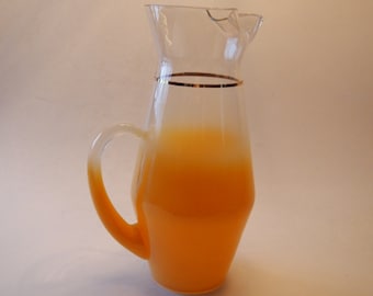 May include: A clear glass pitcher with a gold rim and handle, filled with orange liquid. The pitcher has a classic design, perfect for serving beverages. The orange liquid fills the bottom half of the pitcher.