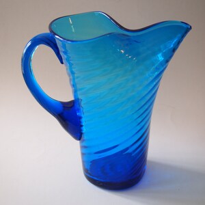 May include: A vibrant blue glass pitcher with a curved handle and a wavy rim. The pitcher features a spiral design and is made of transparent glass, allowing light to pass through. The pitcher is a decorative item.
