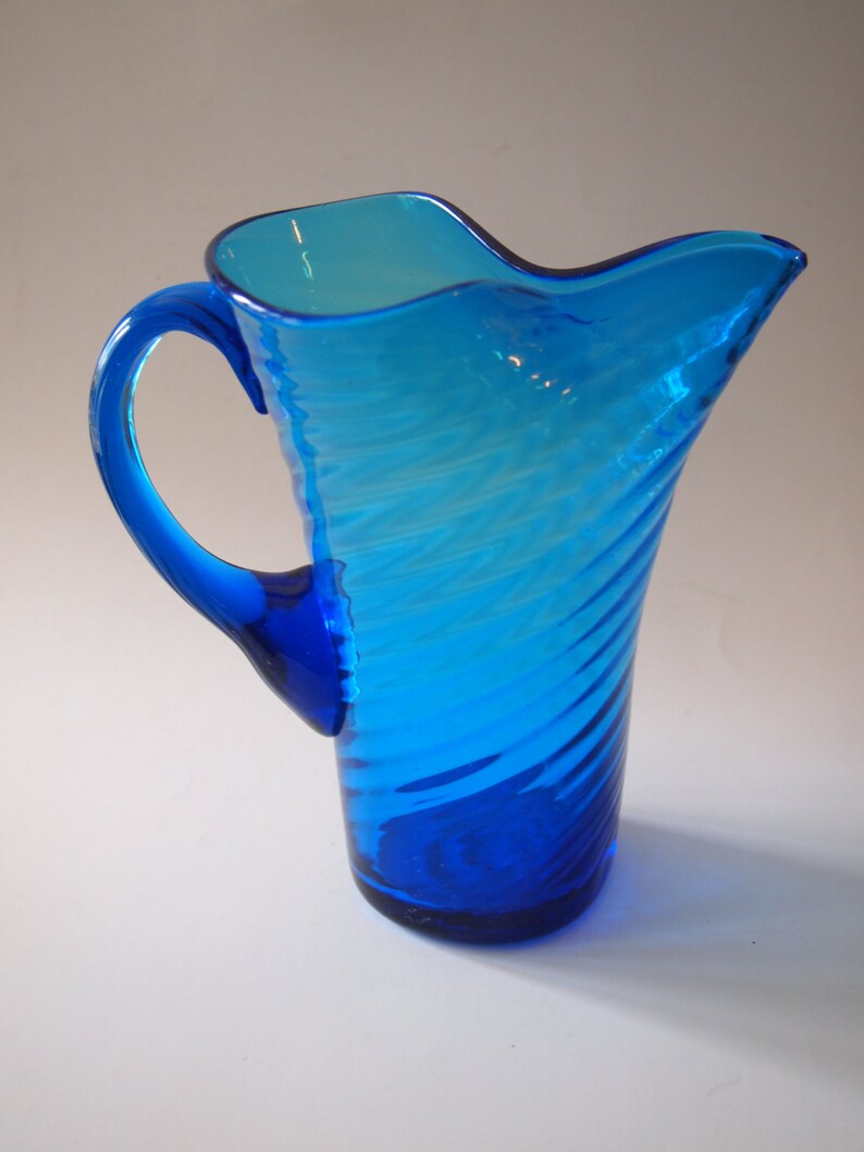 May include: A vibrant blue glass pitcher with a curved handle and a wavy rim. The pitcher features a spiral design and is made of transparent glass, allowing light to pass through. The pitcher is a decorative item.