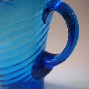 May include: A vibrant blue glass pitcher with a curved handle. The pitcher has a textured, swirled design and a deep blue base. The handle is a darker shade of blue, adding contrast to the overall design. A decorative piece for serving beverages.
