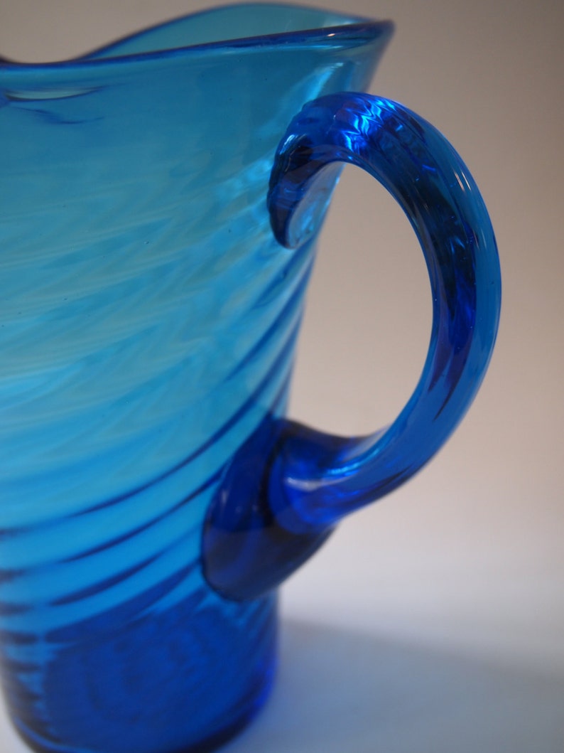 May include: A vibrant blue glass pitcher with a curved handle. The pitcher has a textured, swirled design and a deep blue base. The handle is a darker shade of blue, adding contrast to the overall design. A decorative piece for serving beverages.