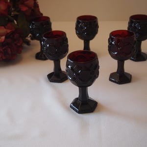 May include: A set of six deep red glass goblets featuring a textured geometric pattern on the bowl and a hexagonal base. Each goblet has a short stem and a rounded cup with intricate embossed detailing, arranged in a cluster on a plain surface.