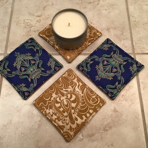 May include: Four square fabric coasters, two with a gold and white paisley design and two with a blue and gold paisley design. A white candle sits on one of the gold coasters.