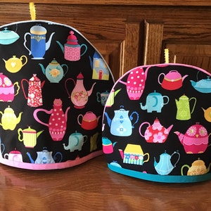 May include: Two teapot cozies with a black background and a colorful pattern of teapots, houses, and animals. The larger cozy has a pink trim and the smaller cozy has a teal trim.