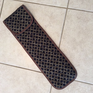 May include: A long, black fabric case with a gold geometric pattern. The case has a brown trim and a flap with a zipper closure.