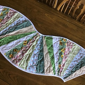 May include: A quilted table runner with a fan-like design. It features various fabrics in shades of green, white, and pink, with floral and sunflower patterns. The runner is approximately 30 inches long and has a white border.