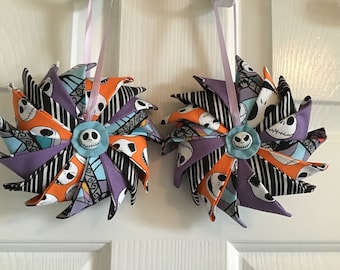 May include: Two handmade fabric ornaments in the shape of pinwheels. The pinwheels are made from a variety of fabrics, including orange, purple, black, and white. The center of each pinwheel features a blue button with a white Jack Skellington face.