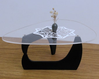 Op de afbeelding: Een driehoekige salontafel van helder glas met een zwarte basis en een wit kantkleedje erop. Een kleine grijze vaas met gedroogde bloemen staat op het kleedje.