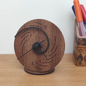 Op de afbeelding: Een houten klok met een uniek spiraalontwerp en zwarte wijzers. De wijzerplaat van de klok heeft de cijfers 1 tot en met 12.