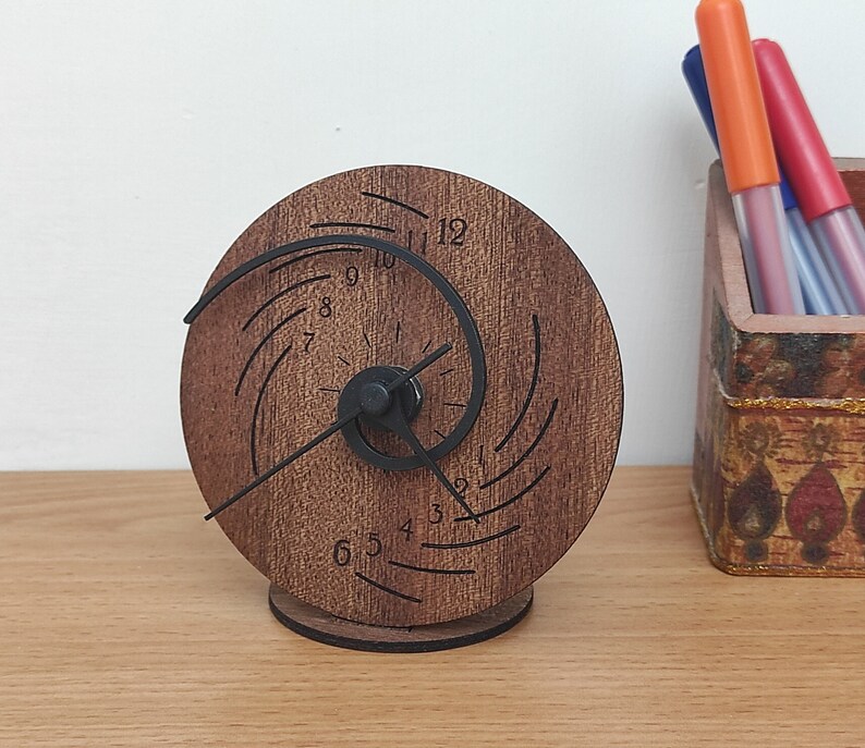 Op de afbeelding: Een houten klok met een uniek spiraalontwerp en zwarte wijzers. De wijzerplaat van de klok heeft de cijfers 1 tot en met 12.
