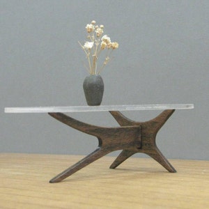 Puede incluir: Una mesa de centro de estilo moderno de mediados de siglo con una tapa de vidrio transparente y una base de madera oscura. La base está hecha de cuatro patas que están conectadas en el medio. Un pequeño jarrón con flores secas se encuentra sobre la mesa.