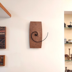 Puede incluir: Un reloj de pared de madera con un diseño en espiral único. La esfera del reloj está hecha de plástico transparente y tiene números negros. Las manecillas del reloj son negras y están unidas a un brazo en espiral que se mueve alrededor de la esfera del reloj.