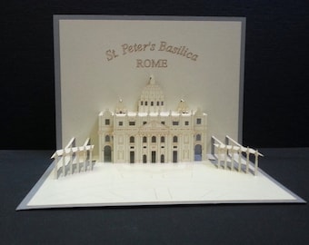 Puede incluir: Una tarjeta pop-up 3D que representa la Basílica de San Pedro en Roma. La tarjeta está hecha de papel y presenta una maqueta detallada de la basílica, incluyendo su cúpula y columnas. La tarjeta está abierta y la basílica es visible en el centro de la tarjeta.