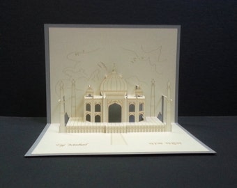 Puede incluir: Una tarjeta de papel pop-up blanca con un modelo 3D de un edificio con una cúpula. La tarjeta está abierta y el edificio es visible. La tarjeta tiene el texto "Taj Mahal" escrito en la parte inferior.
