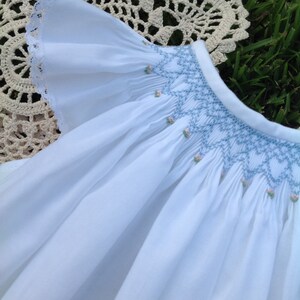 May include: White baby dress with a ruffled sleeve and a smocked bodice. The bodice features light blue embroidery with small pink floral accents. The dress is made of a soft, flowing fabric.