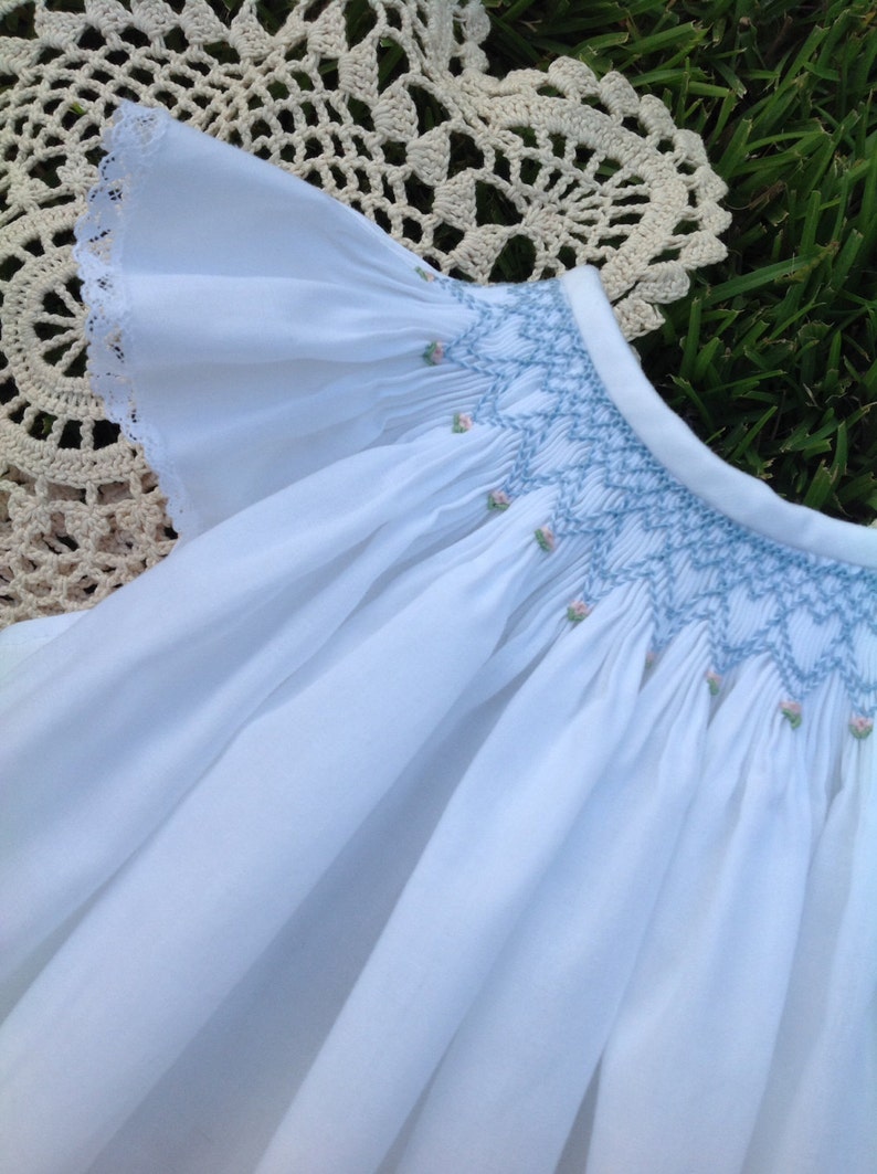 May include: White baby dress with a ruffled sleeve and a smocked bodice. The bodice features light blue embroidery with small pink floral accents. The dress is made of a soft, flowing fabric.
