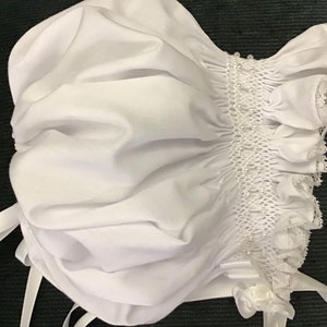May include: White baby bonnet with a ruffled edge and a white satin ribbon. The bonnet has a smocked design with white beads.