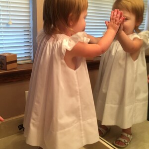 May include: A young child wearing a white dress with ruffled sleeves and white sandals stands in front of a mirror. The child's hands are raised, touching the mirror's surface. The dress is knee-length.