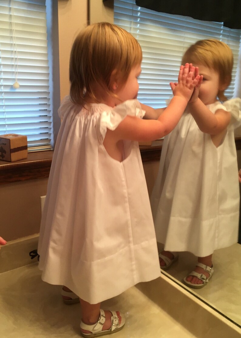 May include: A young child wearing a white dress with ruffled sleeves and white sandals stands in front of a mirror. The child's hands are raised, touching the mirror's surface. The dress is knee-length.