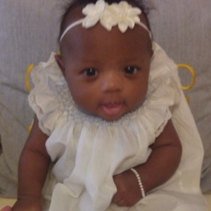 May include: A baby wearing a white dress with a ruffled collar and a white floral headband. A pearl bracelet adorns the baby's wrist. The baby is looking directly at the camera.