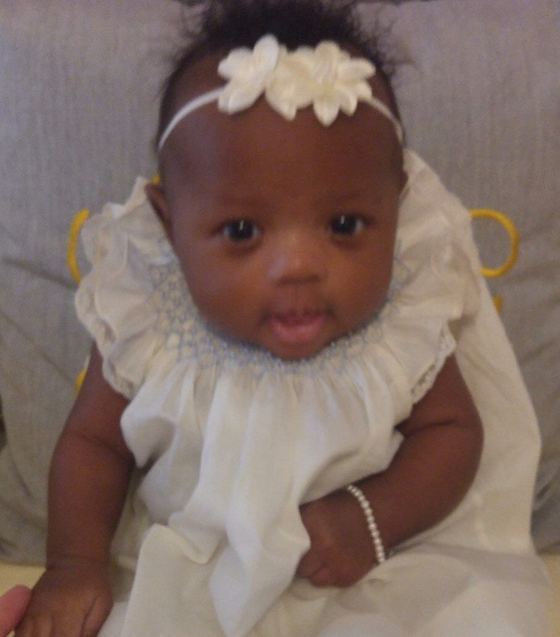 May include: A baby wearing a white dress with a ruffled collar and a white floral headband. A pearl bracelet adorns the baby's wrist. The baby is looking directly at the camera.