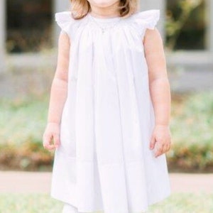 May include: A young child wearing a white dress with ruffled sleeves, white leggings, and white shoes. A large white bow adorns the child's hair. The child is smiling while standing on green grass.