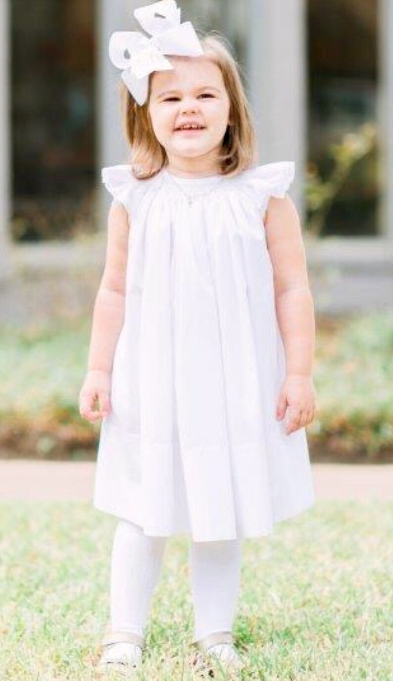 May include: A young child wearing a white dress with ruffled sleeves, white leggings, and white shoes. A large white bow adorns the child's hair. The child is smiling while standing on green grass.