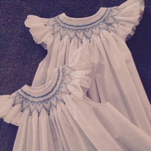 May include: Two white baby dresses with blue embroidered details. The dresses have ruffled sleeves and a smocked neckline. The embroidery features a geometric pattern. The dresses are made of a soft, flowing fabric.