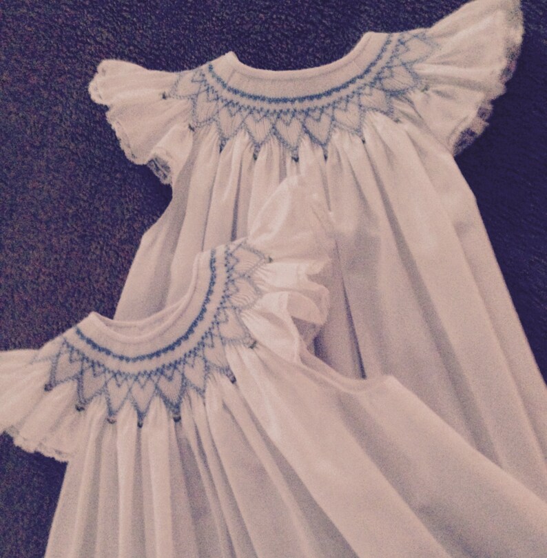 May include: Two white baby dresses with blue embroidered details. The dresses have ruffled sleeves and a smocked neckline. The embroidery features a geometric pattern. The dresses are made of a soft, flowing fabric.