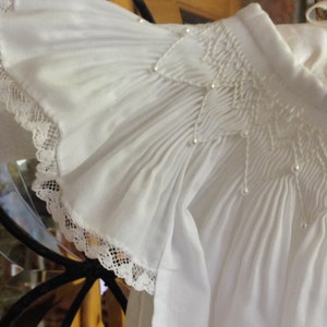 May include: Close-up of a white, ruffled garment with delicate lace trim. The fabric is gathered and smocked, with small pearl accents. The sleeve features a ruffled edge. The garment appears to be a formal top or dress.