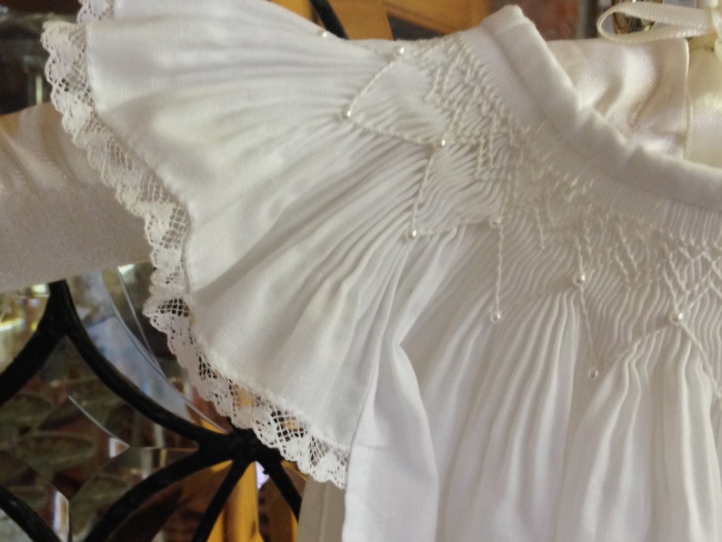 May include: Close-up of a white, ruffled garment with delicate lace trim. The fabric is gathered and smocked, with small pearl accents. The sleeve features a ruffled edge. The garment appears to be a formal top or dress.