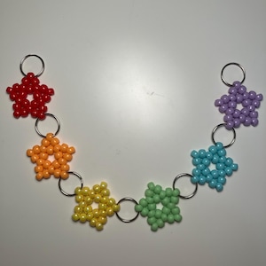 May include: A chain of colorful star-shaped beaded keychains in a rainbow of colors: red, orange, yellow, green, blue, and purple. Each star is connected by a silver ring and has a silver keyring attached.
