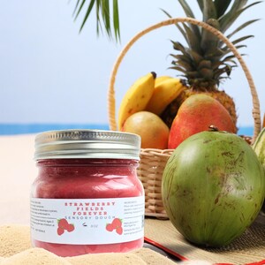 May include: A glass jar of red strawberry-flavored sensory dough with a white lid. The jar is labeled "Strawberry Fields Forever Sensory Dough" and has a picture of a strawberry on the label. The jar is sitting on a beach with a basket of fruit in the background.
