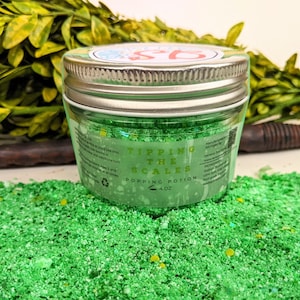 May include: A small glass jar filled with green bath salts and glitter. The jar has a silver lid and a label that reads "Tipping the Scales Popping Potion 4 oz".