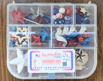 May include: A clear plastic storage box with 9 compartments filled with red, white, and blue craft supplies. The box has a label that says "Sensory Box Family Sensory Box An invitation to play Sweet Land of Liberty".