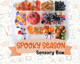 May include: A clear plastic divided storage container filled with Halloween-themed items including orange and clear glass gems, gold pipe cleaners, black stones, plastic spiders, colorful pom-poms, googly eyes, and a textured pumpkin. Text reads SPOOKY SEASON Sensory Box.