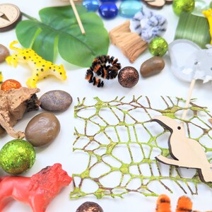 May include: A collection of jungle-themed toys and objects, including a yellow leopard, a brown toucan, a green leafy plant, a brown wooden log, a compass, a grey elephant, a yellow giraffe, a brown wooden stick, a green net, and brown and orange rocks.