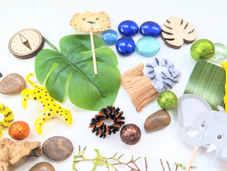 May include: A collection of jungle-themed toys and decorations, including a leopard figurine, a wooden elephant, a green leaf, a compass, and blue and green glass gems.