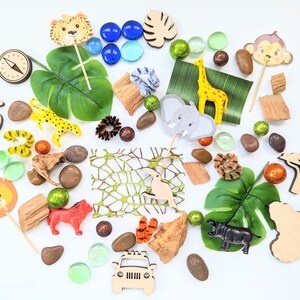 May include: A collection of jungle animal themed toys and decorations, including a lion, giraffe, elephant, monkey, leopard, and a toucan. There are also green leaves, brown rocks, and a wooden jeep.