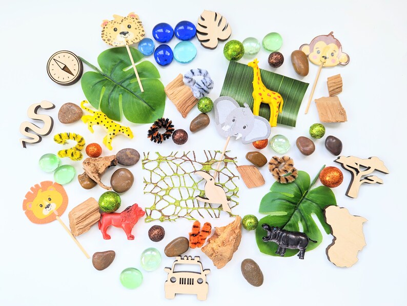 May include: A collection of jungle animal themed toys and decorations, including a lion, giraffe, elephant, monkey, leopard, and a toucan. There are also green leaves, brown rocks, and a wooden jeep.