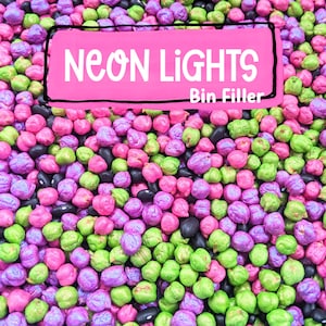 May include: A collection of small, round, dyed dried chickpeas in vibrant neon shades of pink, purple, and lime green, mixed with solid black beans. A pink rectangular label with a black border displays the text NEON LIGHTS Bin Filler.