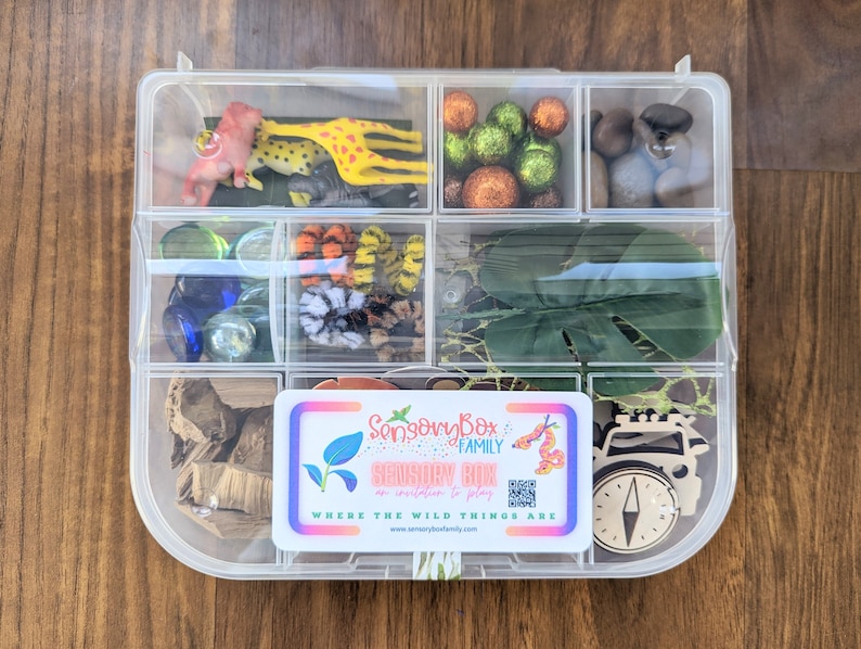 May include: A clear plastic storage box with compartments filled with various sensory items, including small plastic animals, colorful beads, glass marbles, and a wooden compass. The box has a label that reads "Sensory Box Family Sensory Bin Where the Wild Things Are".