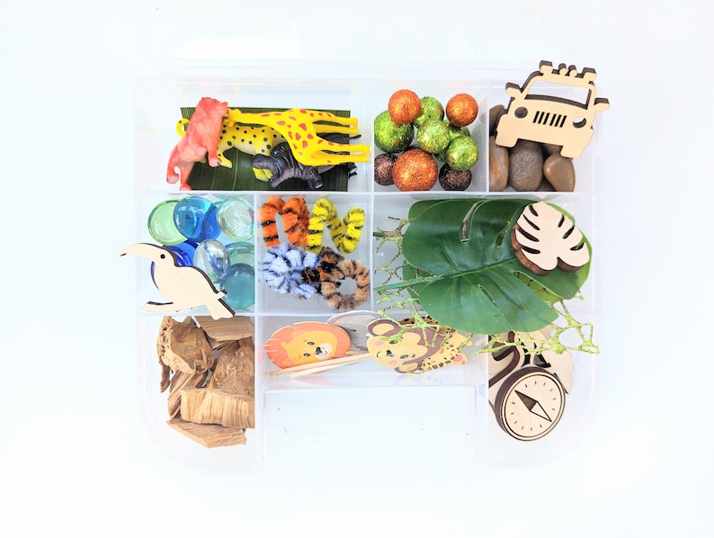 May include: A clear plastic storage box filled with various jungle-themed toys and craft supplies. The box includes a wooden jeep, a toucan, a compass, a tiger, a giraffe, a monkey, leaves, and colorful glass gems.