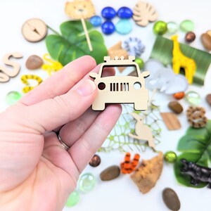 May include: Wooden toy jeep cutout, a part of a larger set of safari-themed toys. The jeep is light brown and has a simple design with a grill, headlights, and a roof rack. The jeep is surrounded by other safari-themed toys, including a giraffe, a monkey, a snake, and a compass.